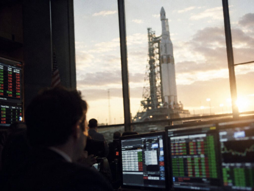 Heavy-lift rocket on a launch pad seen through the window of a financial trading floor
