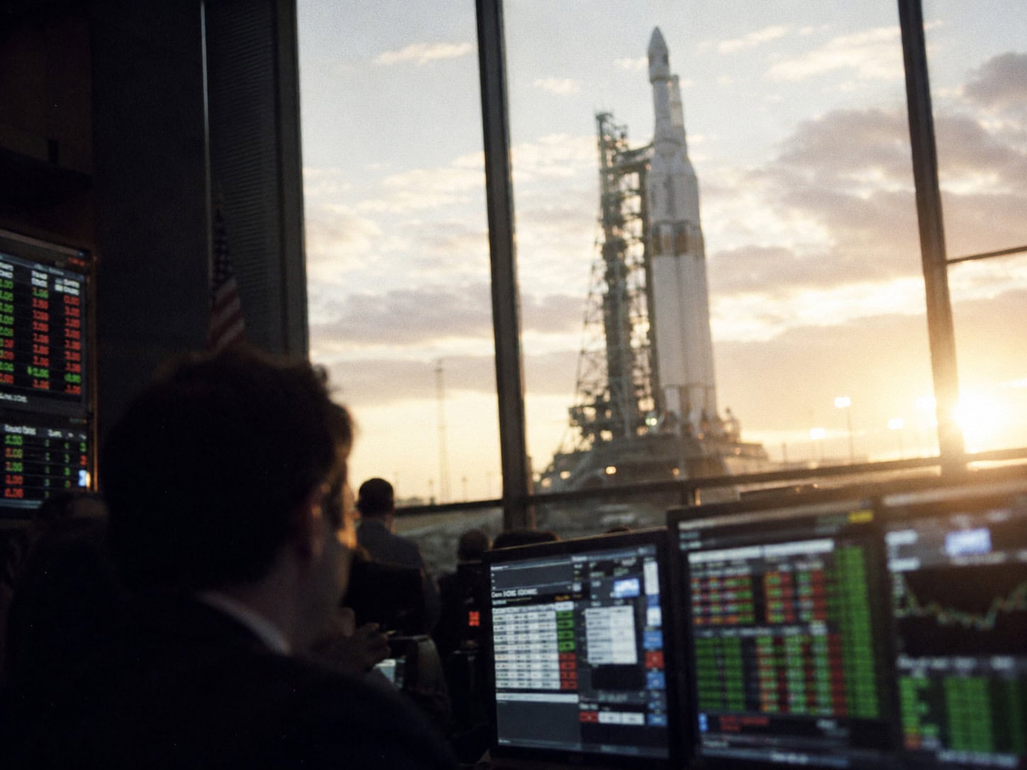 Heavy-lift rocket on a launch pad seen through the window of a financial trading floor