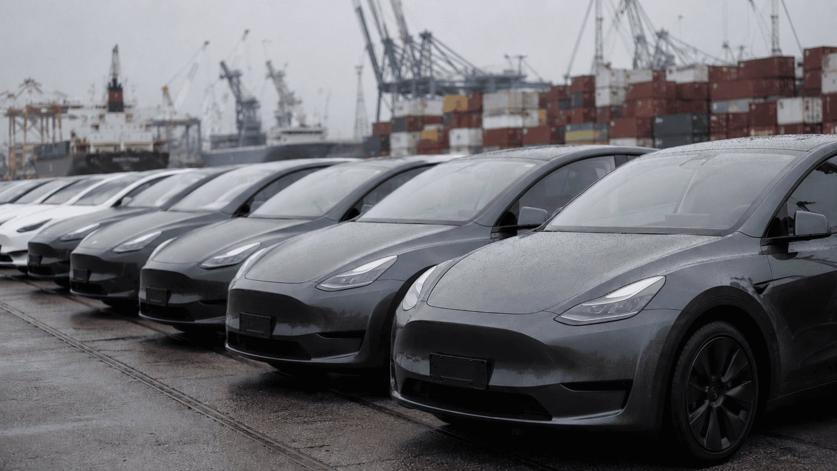 Rows of new electric cars parked at a European shipping port with cranes and stacked containers in the background under overcast sky.
