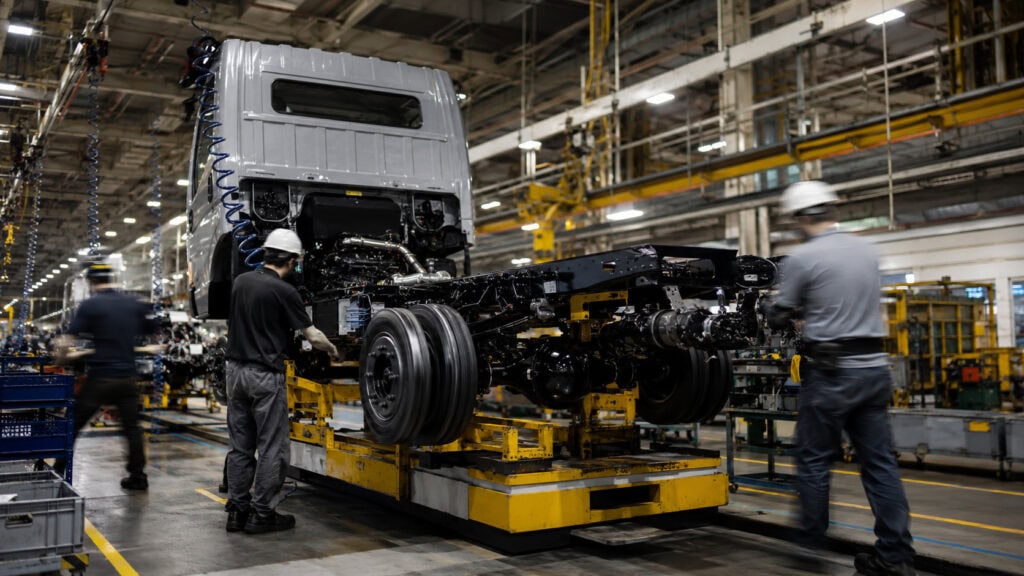Workers in safety gear assembling a vehicle on a yellow lift rig inside a brightly lit automotive factory.