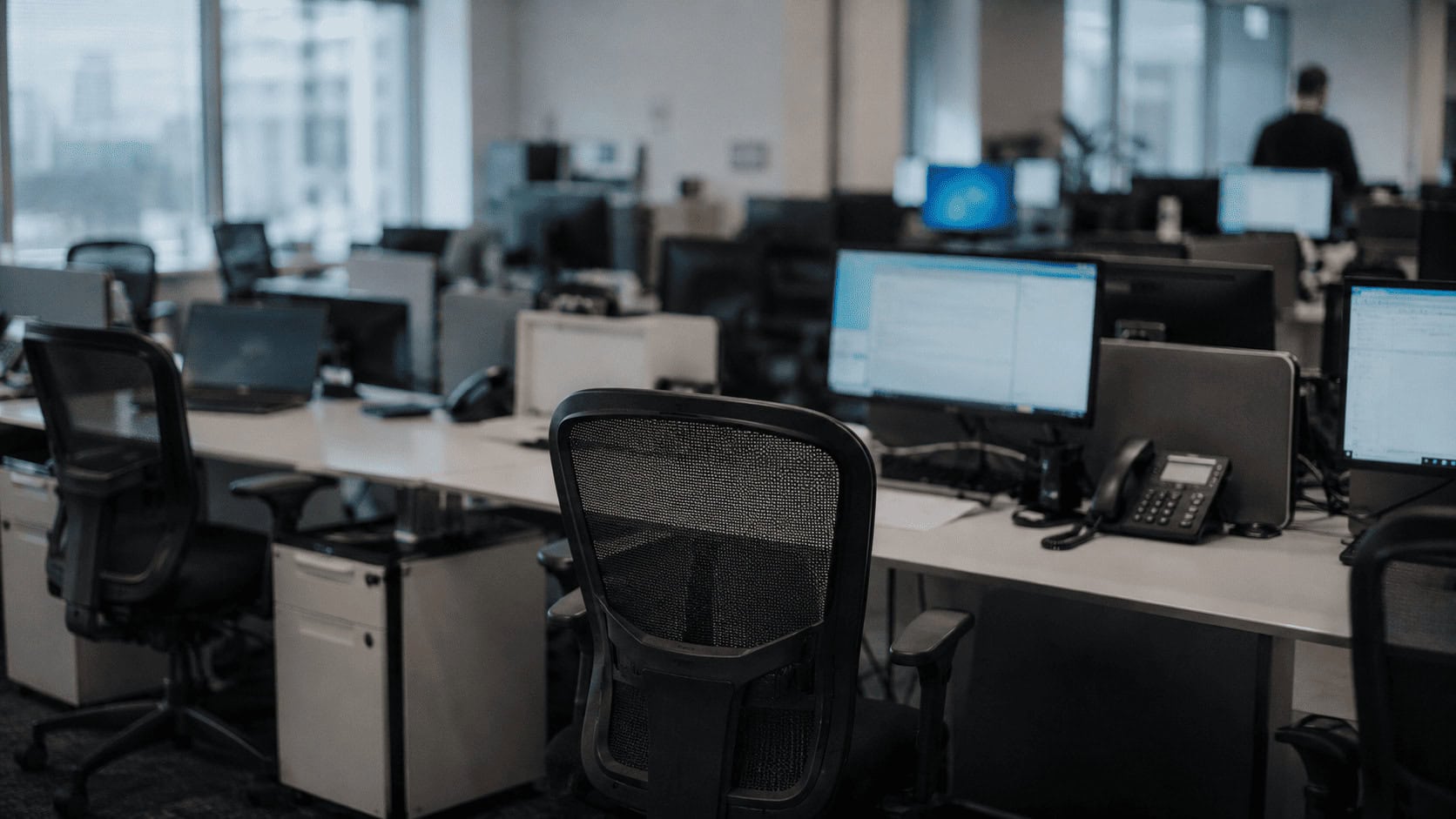 Empty desks and unoccupied chairs in a modern open-plan office with monitors switched on.
