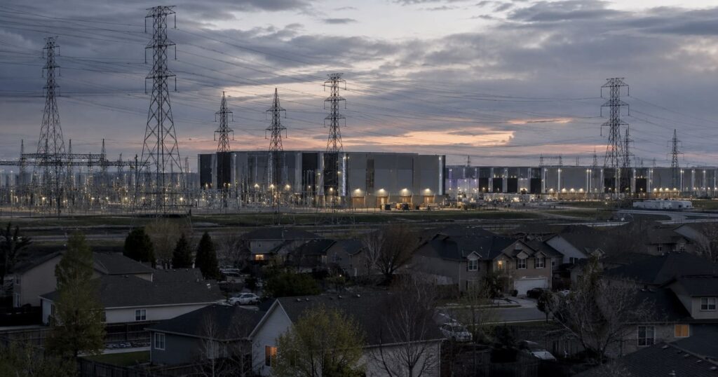 A data center campus beside high-voltage transmission towers at dusk, with suburban homes in the foreground, illustrating AI power demand straining the electric grid.