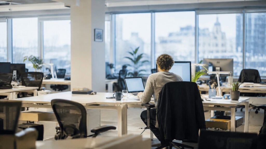 A lone software engineer works at a lit desk in a darkened open-plan office, surrounded by empty workstations.