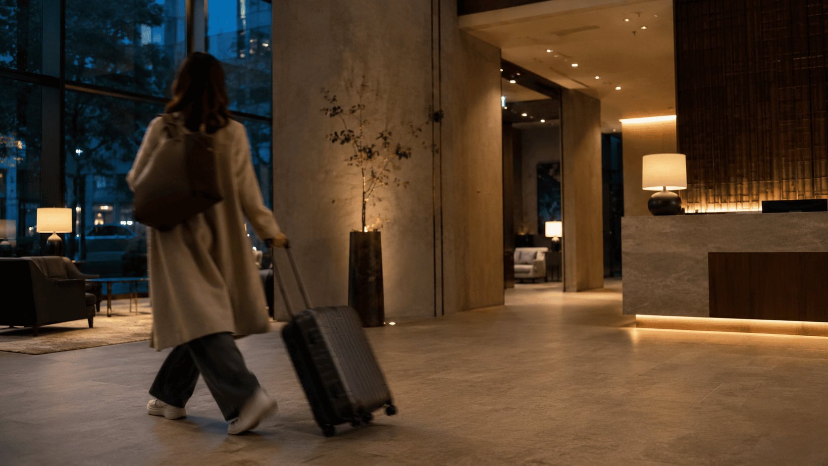 A traveler with a roller suitcase walks through the warmly lit lobby of a modern boutique hotel at dusk.
