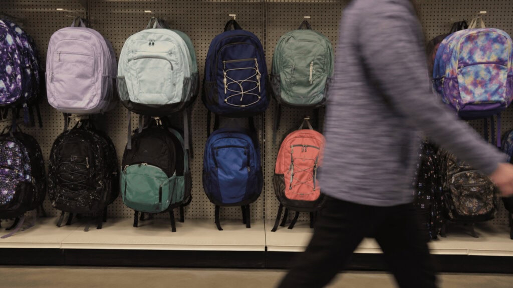 A wall of colorful backpacks on store pegboard hooks with a shopper walking past in the foreground.