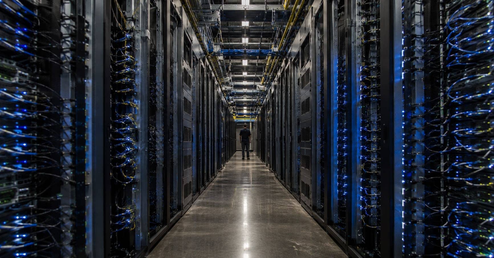 Wide view down the aisle of a modern data center lined with illuminated server racks, illustrating big tech profitability and operating margins.