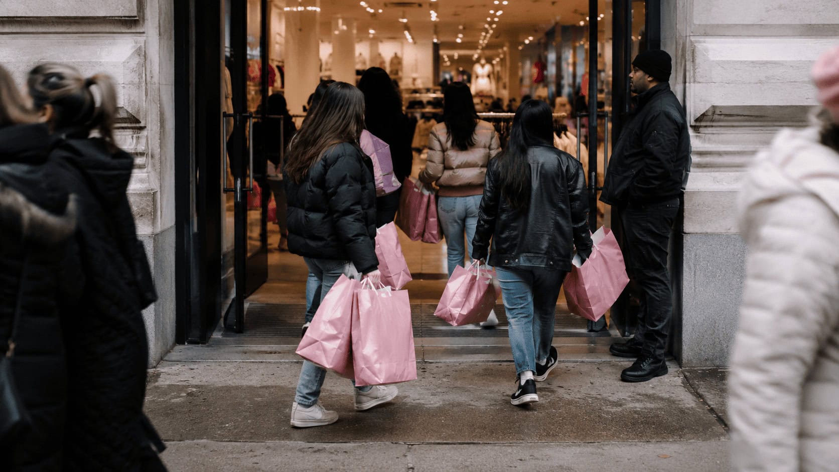 Young shoppers carrying pink shopping bags exit a busy clothing store as a security guard stands near the entrance.