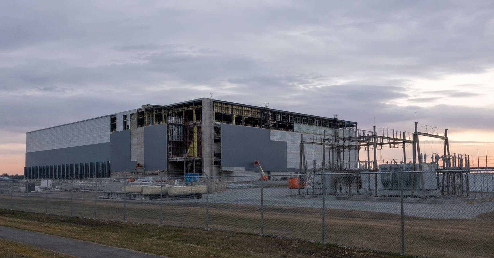 A data center under construction at dusk behind a chain-link fence with substation equipment nearby.
