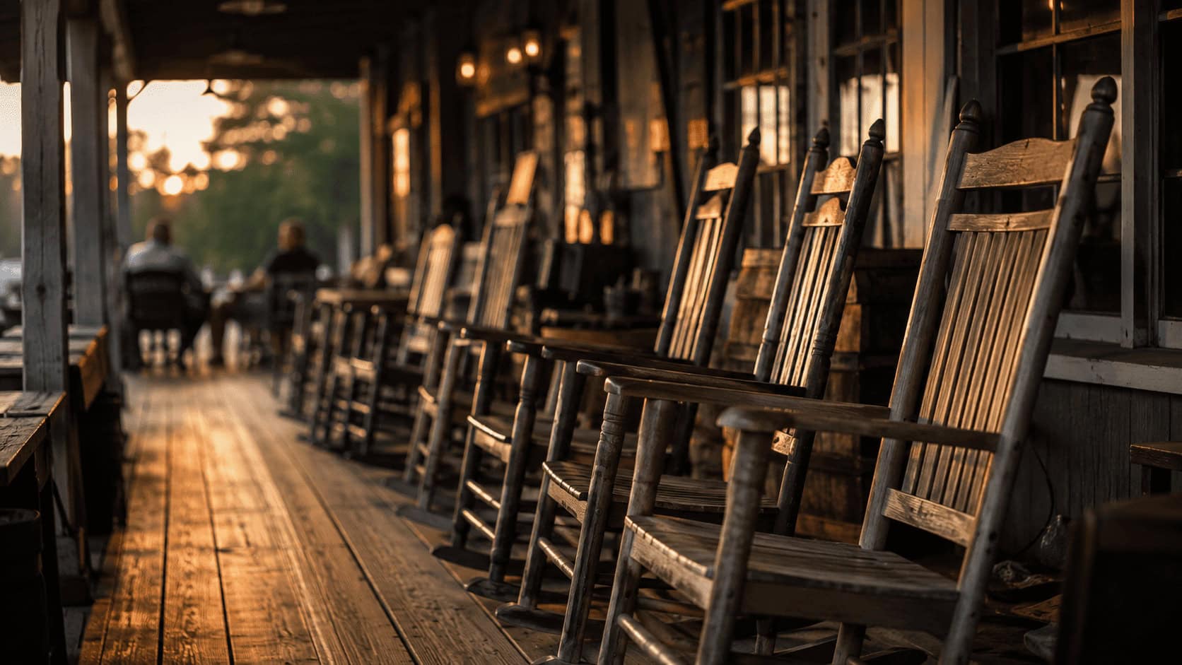 Row of empty wooden rocking chairs along a covered country-store porch at golden hour, with blurred patrons in the background.