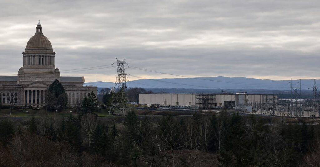 A state capitol building at dusk with a data center, substation, and high-voltage power lines in the distance.