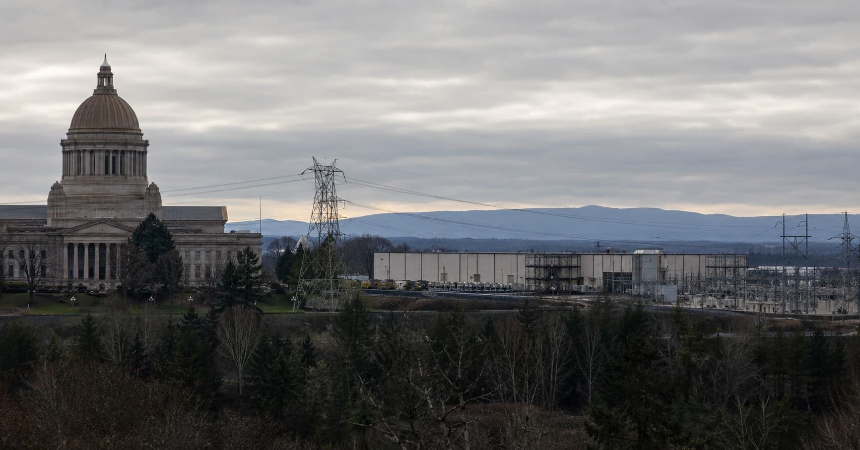 A state capitol building at dusk with a data center, substation, and high-voltage power lines in the distance.