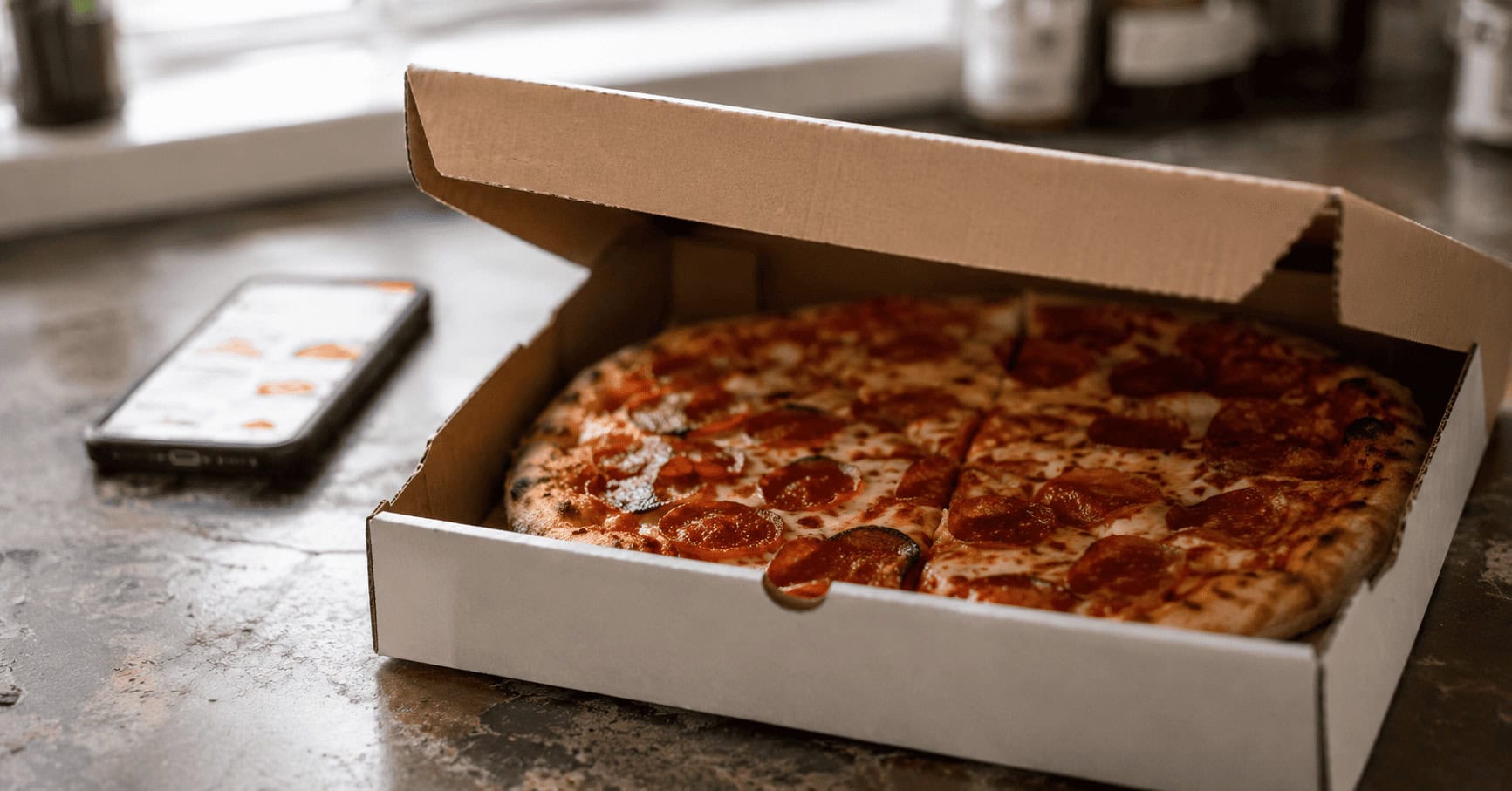 A partially open pizza delivery box holding a cooling pepperoni pizza on a kitchen counter, with a food-delivery app visible on a blurred phone in the background.