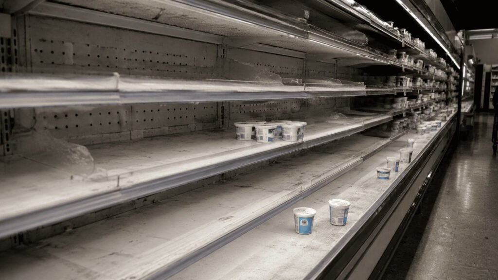 Mostly empty refrigerated grocery store shelf with a few remaining tubs of cottage cheese under fluorescent lighting.