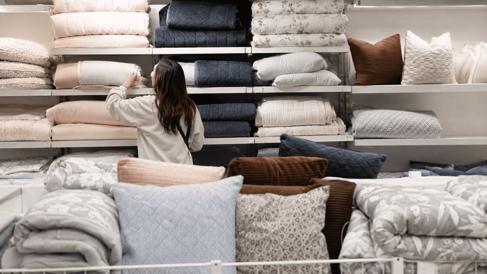 Shopper viewed from behind browsing folded bedding and pillows on shelves in a bright retail home aisle.