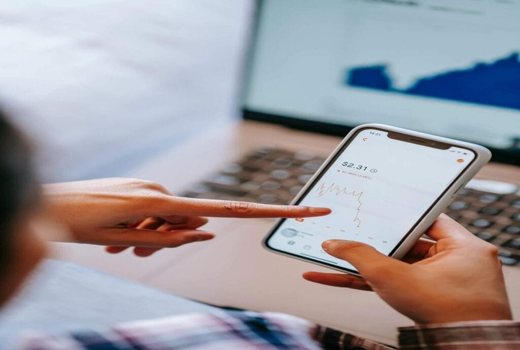 Person analyzing financial data on smartphone with laptop in background.