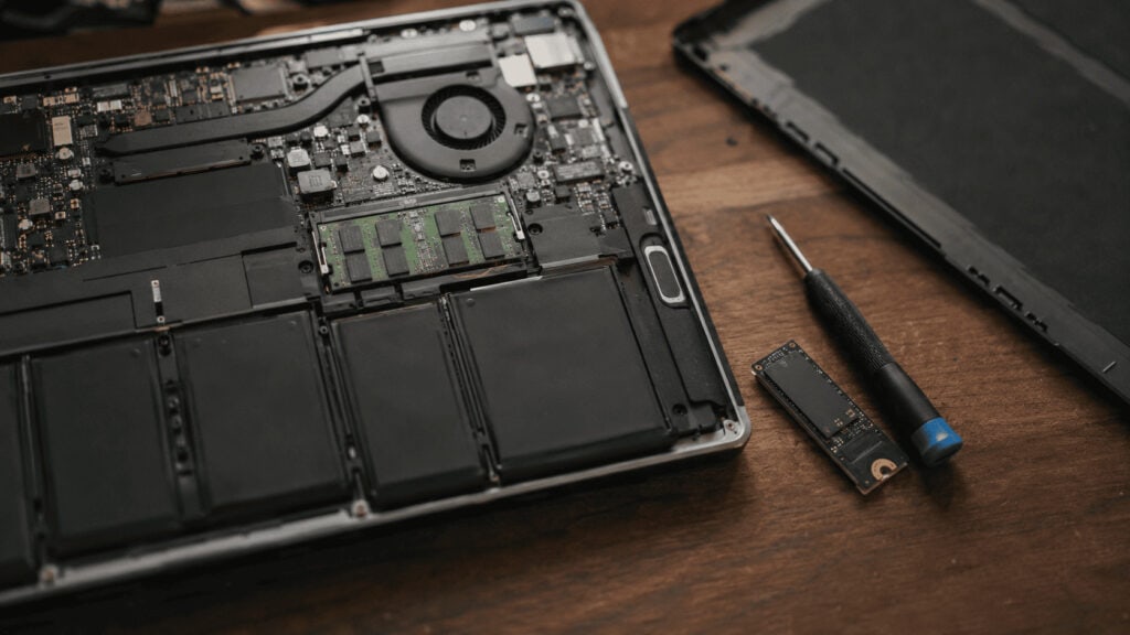 Open laptop mid-repair on a workbench with the back panel removed, exposing RAM modules and battery beside a screwdriver, illustrating the tech repair economy driven by rising 2026 prices