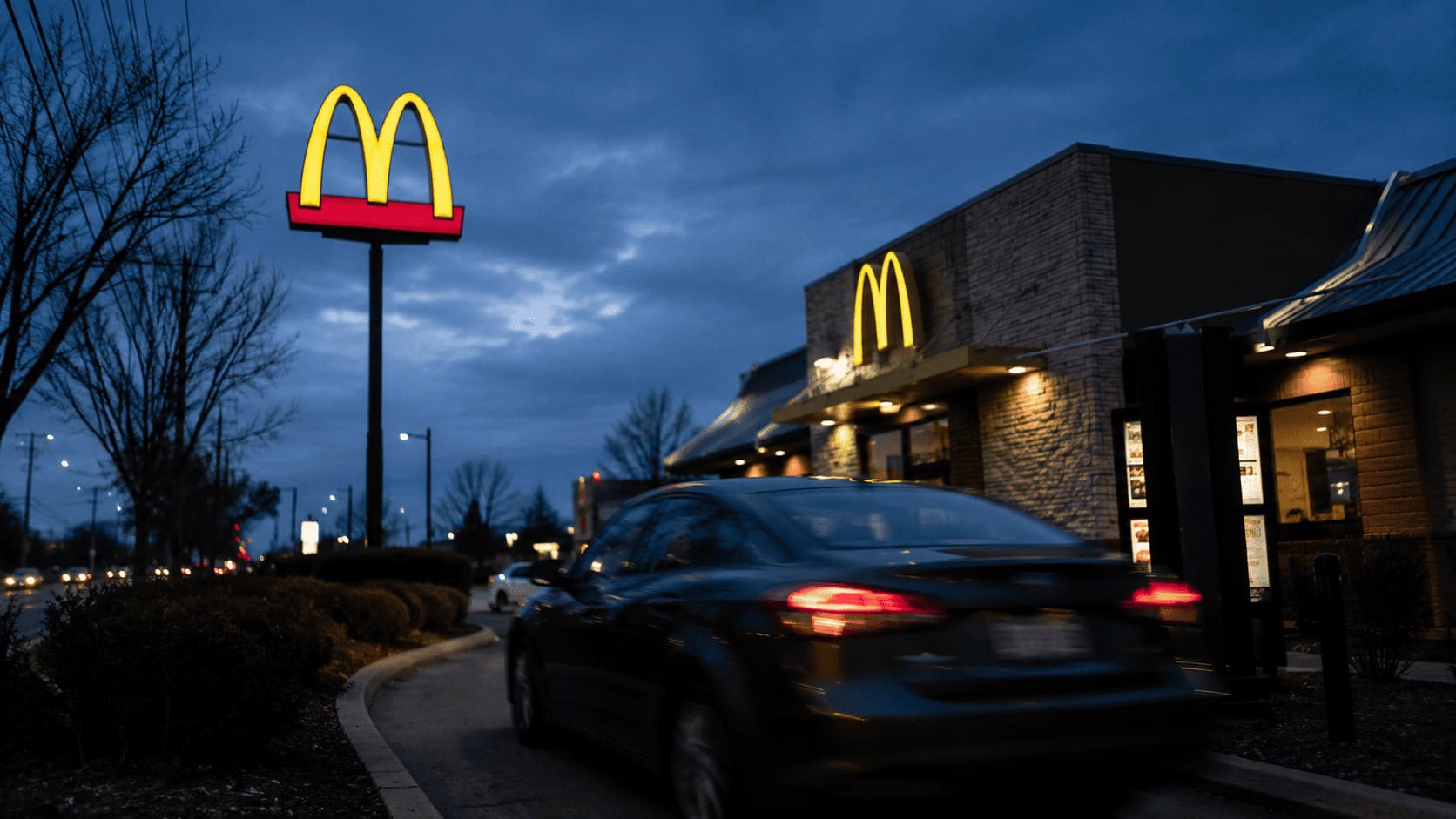 A McDonald's drive-thru exterior at dusk with the golden arches sign illuminated against a blue sky, a car moving through the lane.