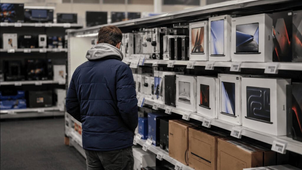 Shopper viewed from behind browsing boxed laptops and game consoles on retail shelves in a brightly lit electronics aisle.