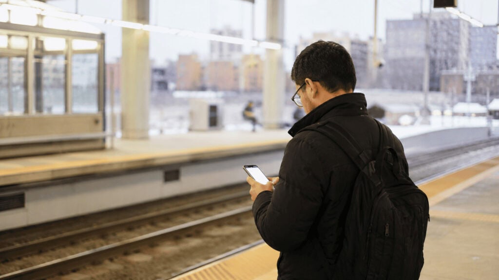 A commuter looks at a glowing smartphone screen on a dimly lit subway platform at dusk.