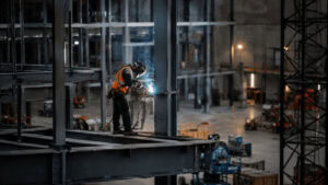 A welder in safety gear works on steel framework at an industrial construction site.