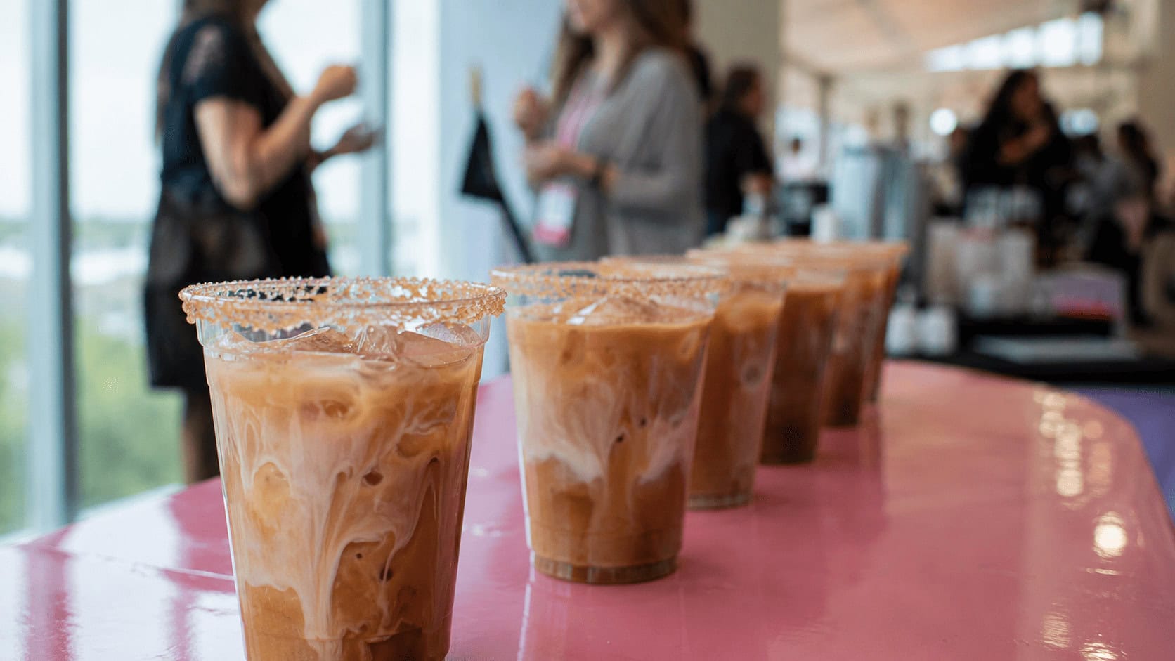 Row of iced oat milk coffee drinks in clear cups with sugar-rimmed edges on a pink table at a beverage event.