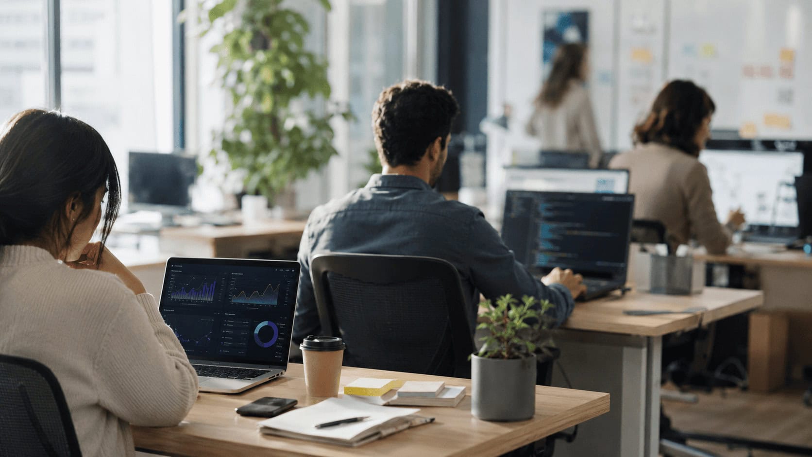 Office professionals working at laptops in an open-plan workspace with screens showing dashboards and code, soft daylight.