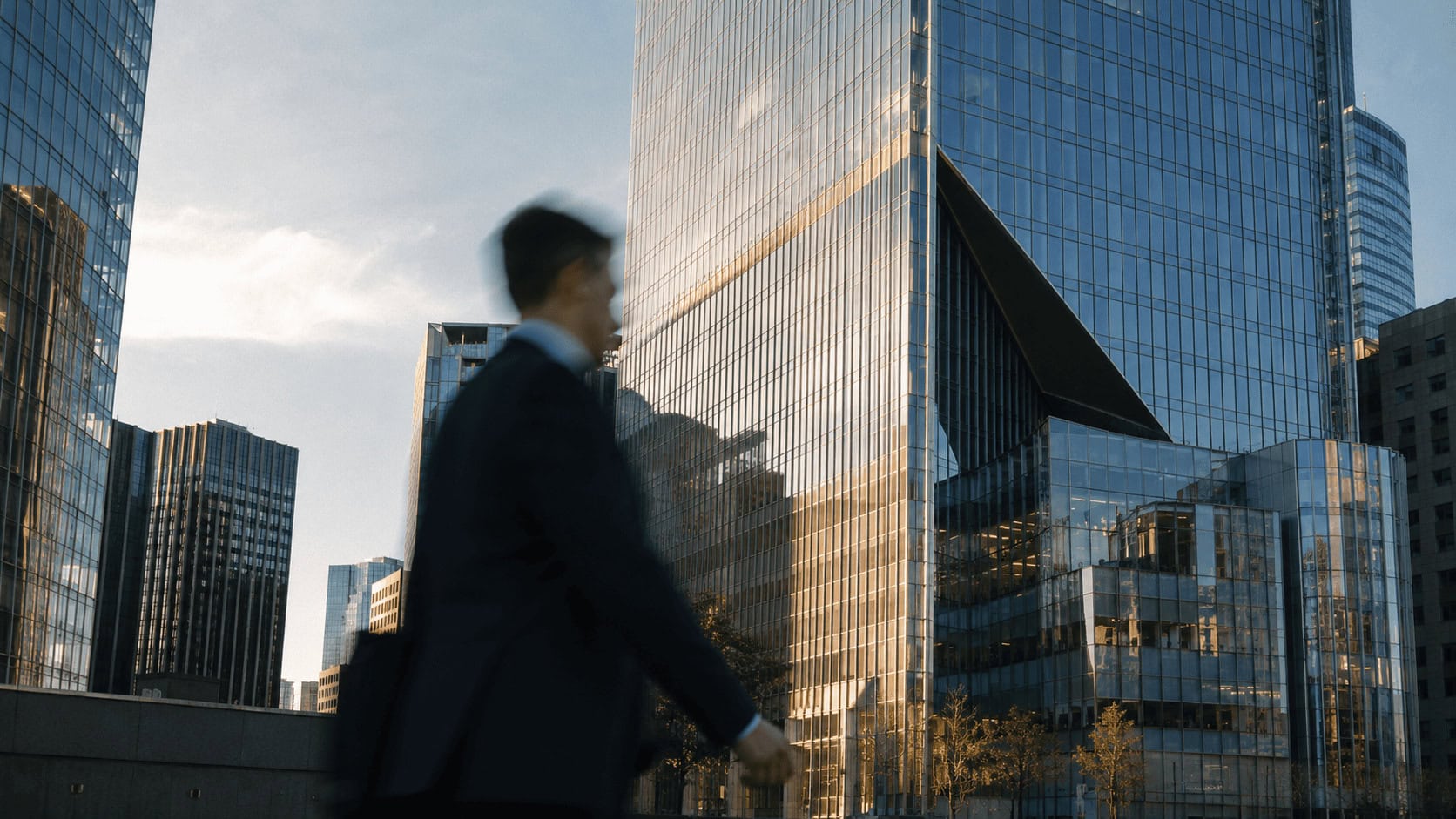 A business professional walks past a glass corporate office tower in a financial district at golden hour.