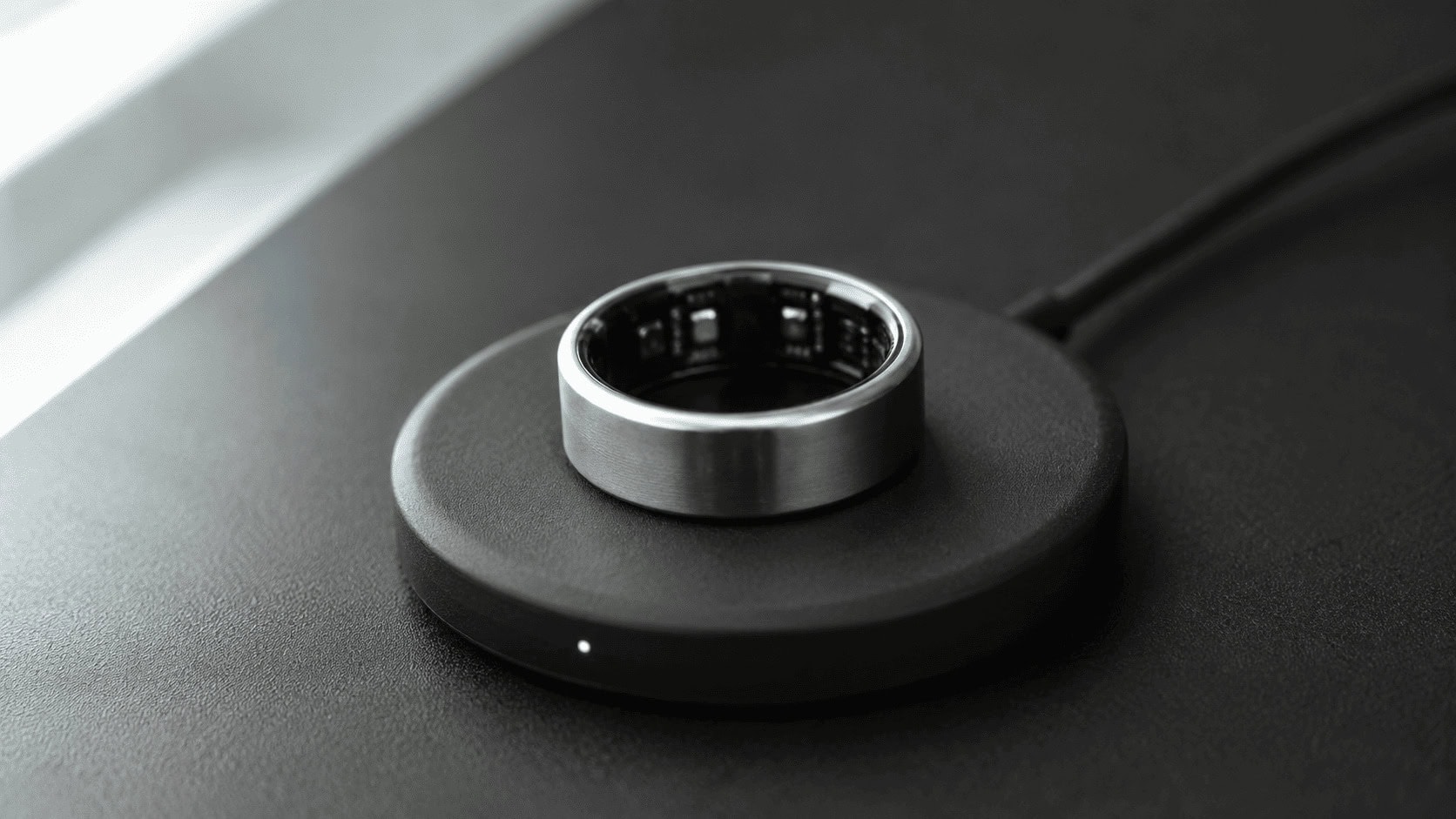 Titanium smart ring resting on a circular wireless charging plate on a charcoal surface