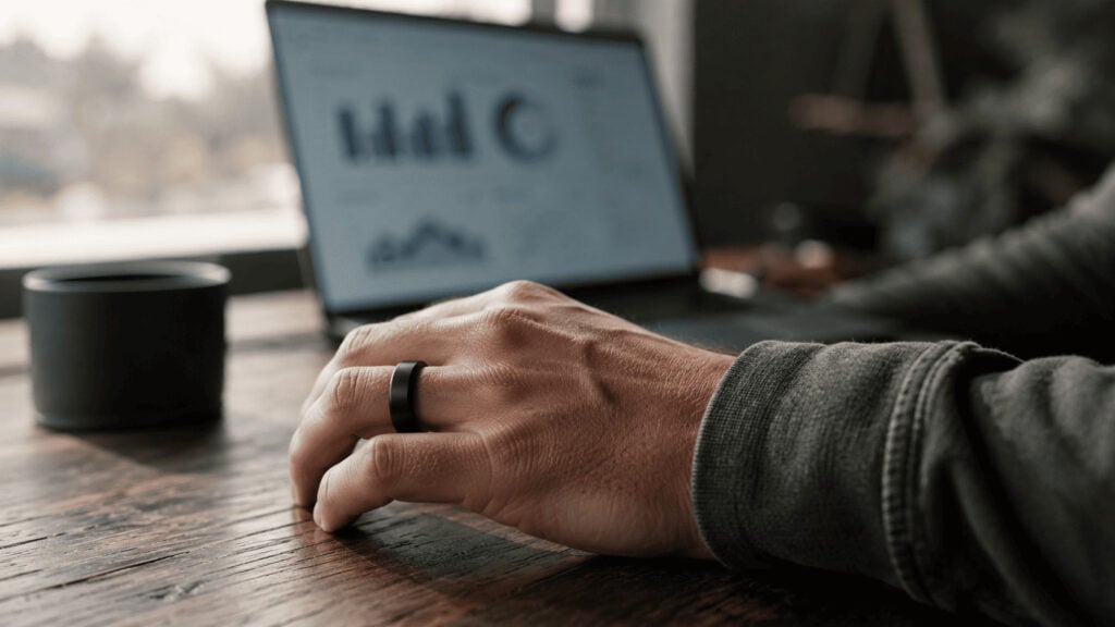 A hand wearing a black smart ring rests on a desk beside a laptop displaying blurred health data charts.