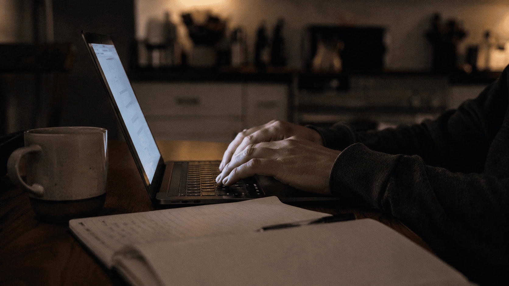 A person works on a laptop at a kitchen table at night, lit mainly by the screen, with a notebook and coffee mug nearby.