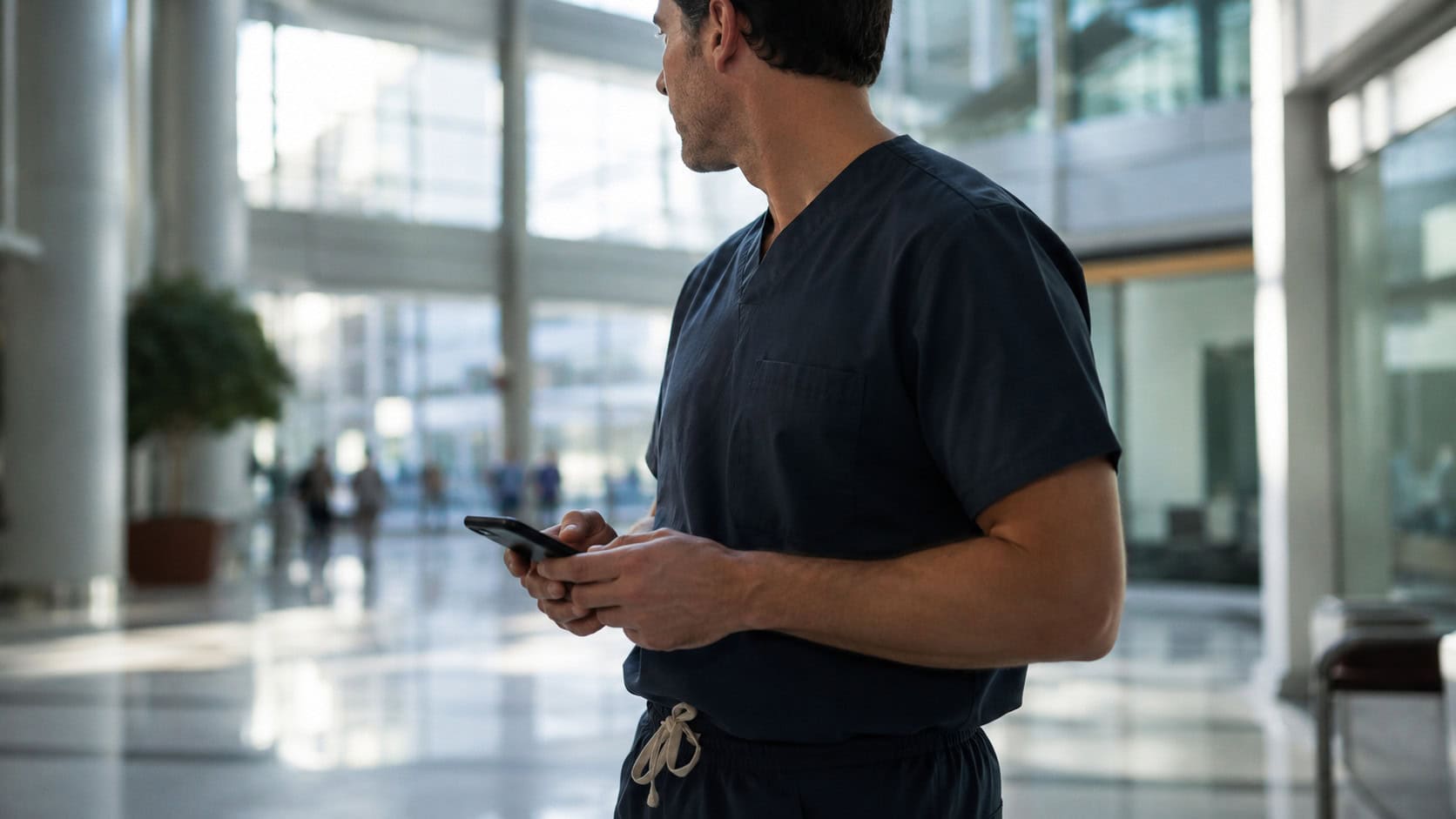 A doctor in dark scrubs holds a smartphone while standing in a sunlit hospital atrium.