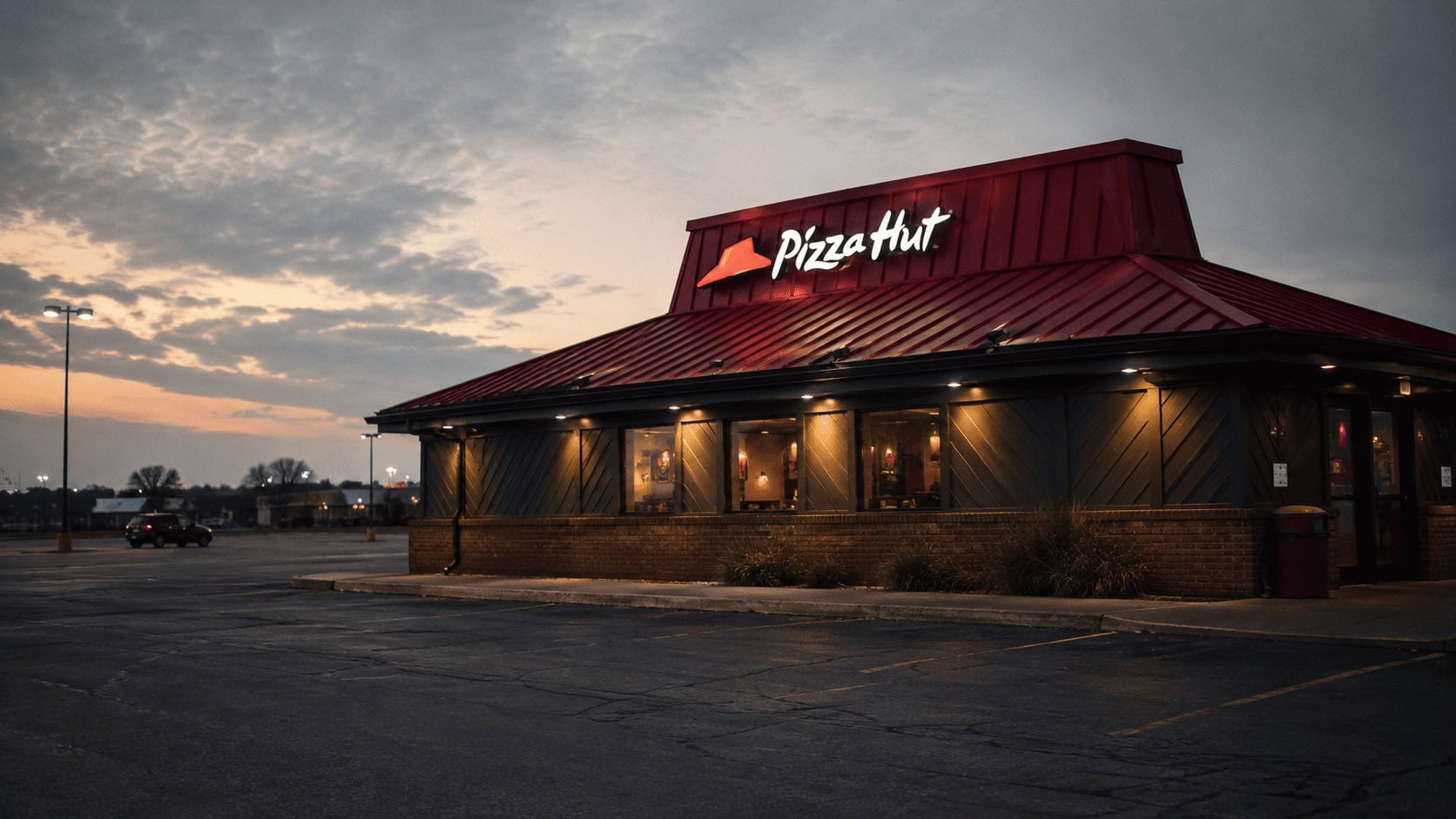 A Pizza Hut restaurant exterior with its red roof at dusk, an empty parking lot in the foreground.