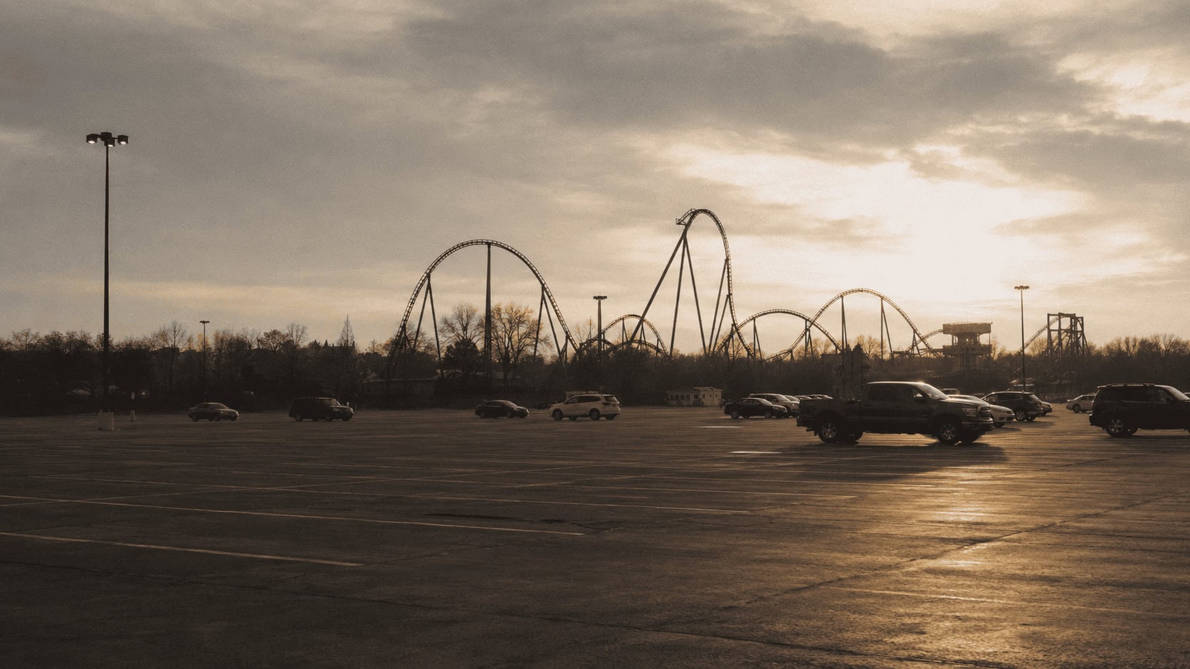 A nearly empty amusement park parking lot at sunset with a roller coaster track visible in the distance under an overcast sky.