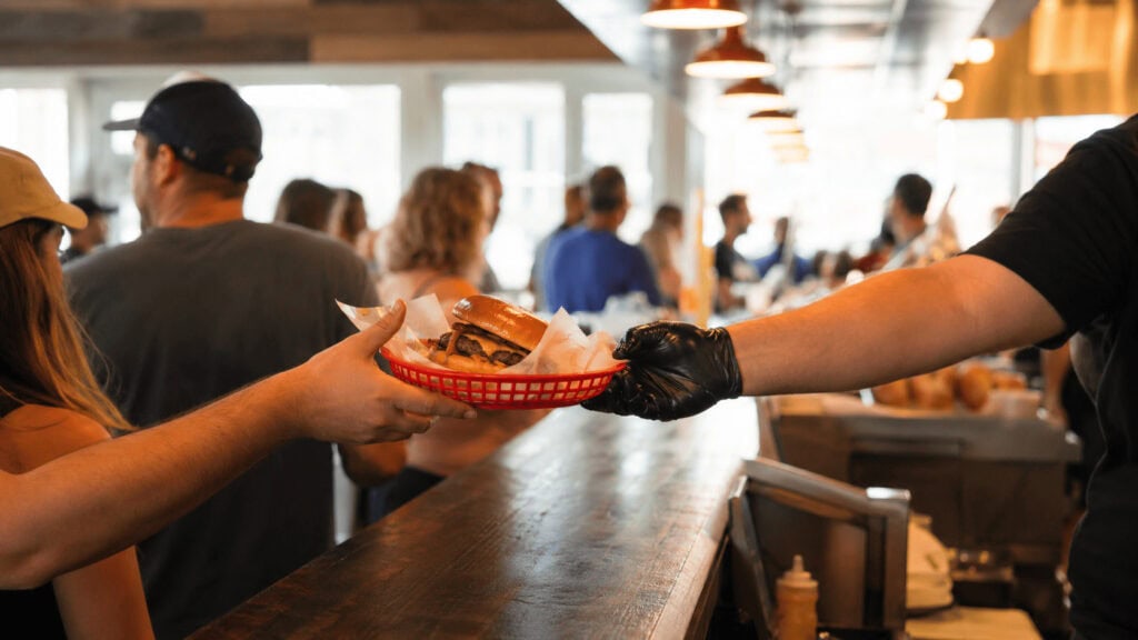 Staff handing fresh burgers in red baskets across a busy restaurant counter as customers wait in line.