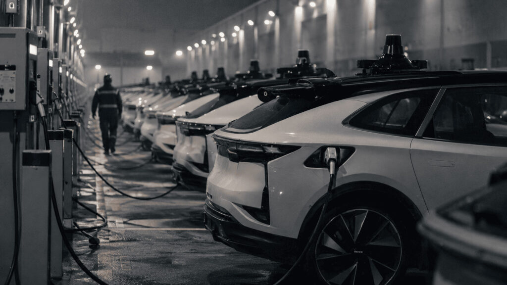 Row of driverless electric taxis parked and charging inside a depot at night, with a worker walking between the vehicles.
