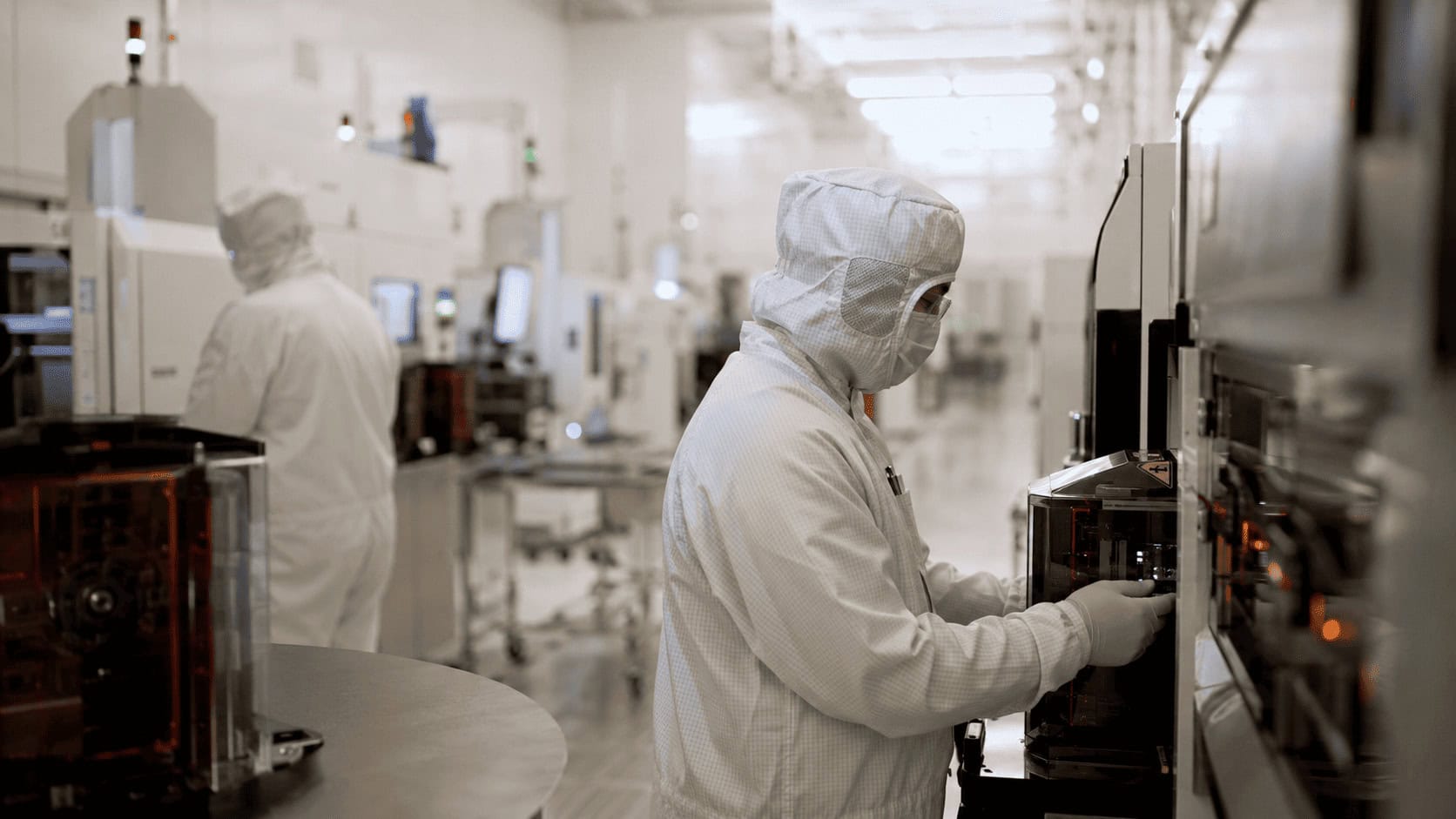 Technicians in white protective suits working inside a brightly lit semiconductor fabrication clean room.