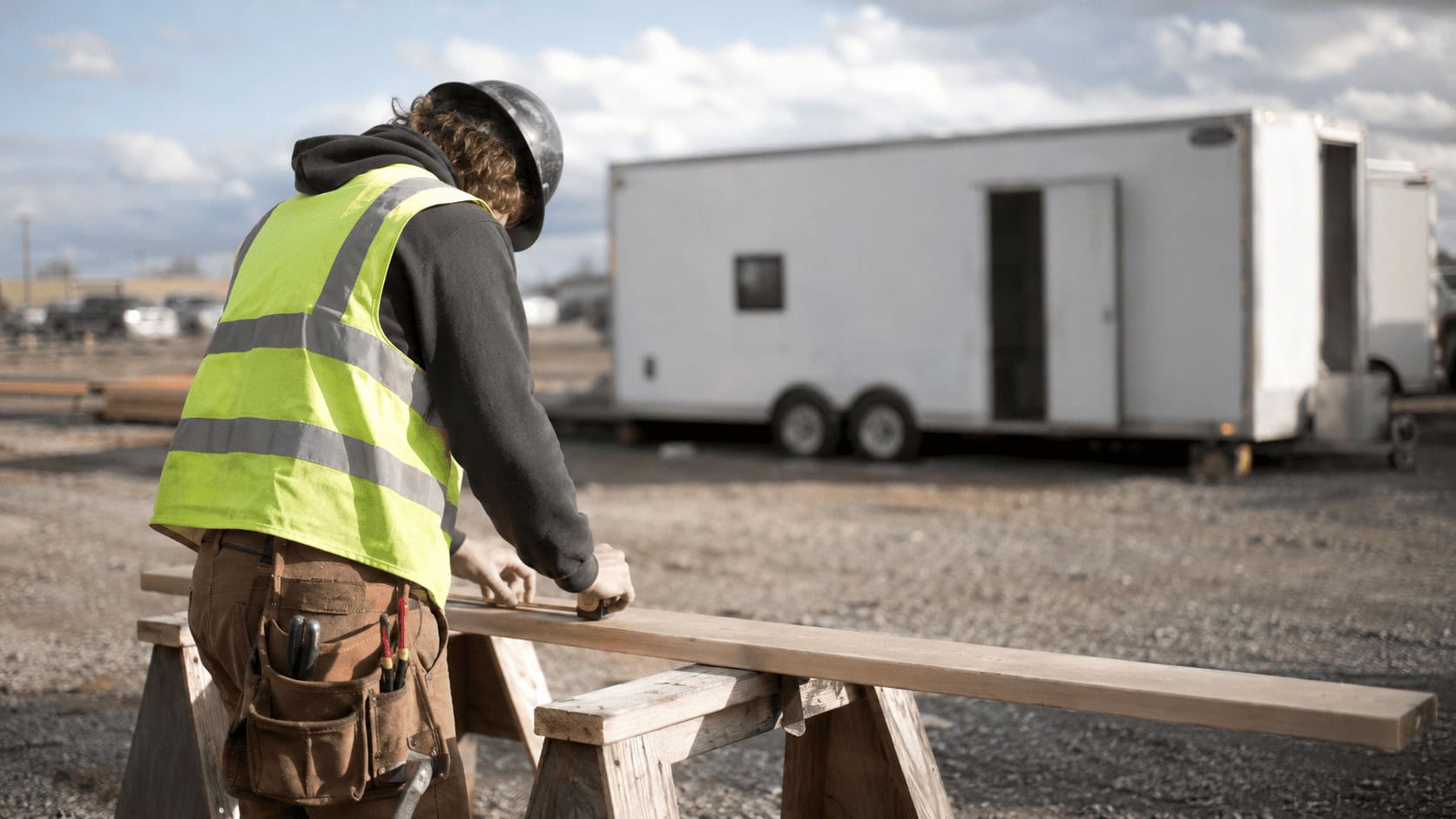 Apprentice in a safety vest cutting lumber at a sawhorse beside a mobile training trailer on a gravel jobsite.