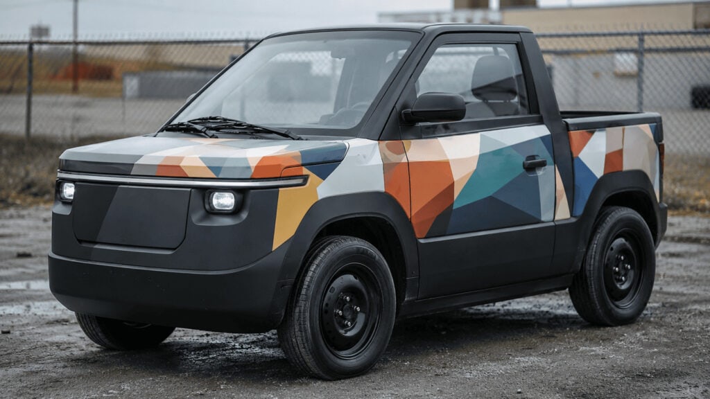 Compact two-seat electric pickup truck with a colorful vinyl wrap parked in an industrial lot.