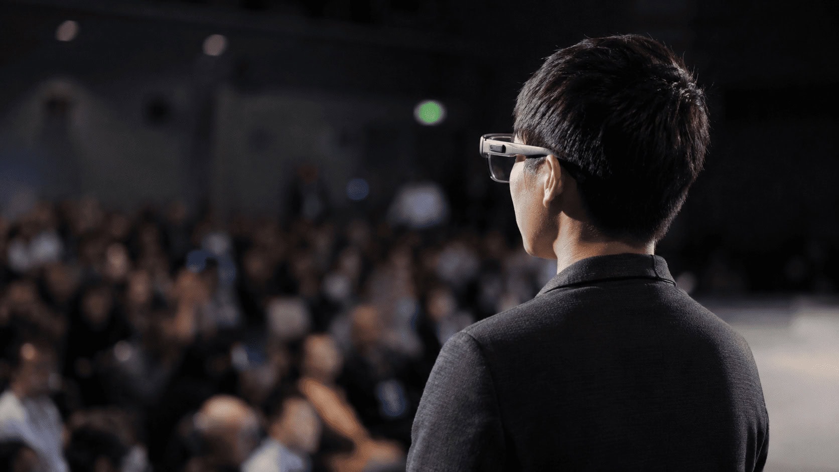 Person wearing transparent augmented reality glasses on a lit stage at a technology expo.