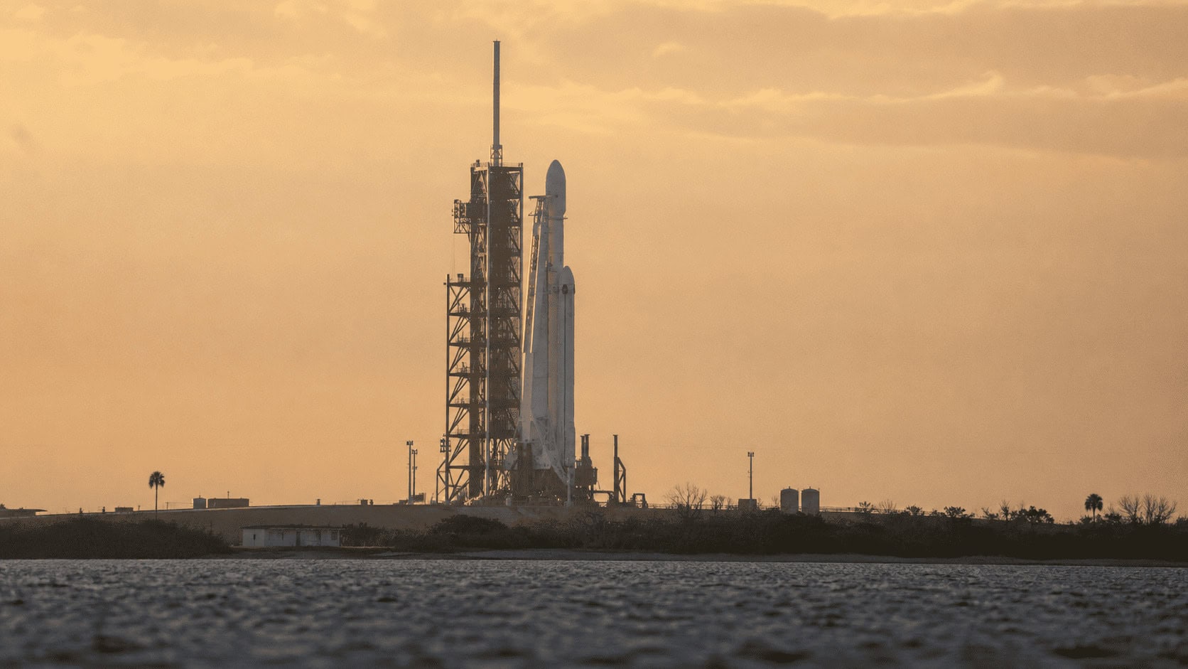 A white rocket stands on a coastal launch pad at dawn under soft sunlight with sea mist in the background.