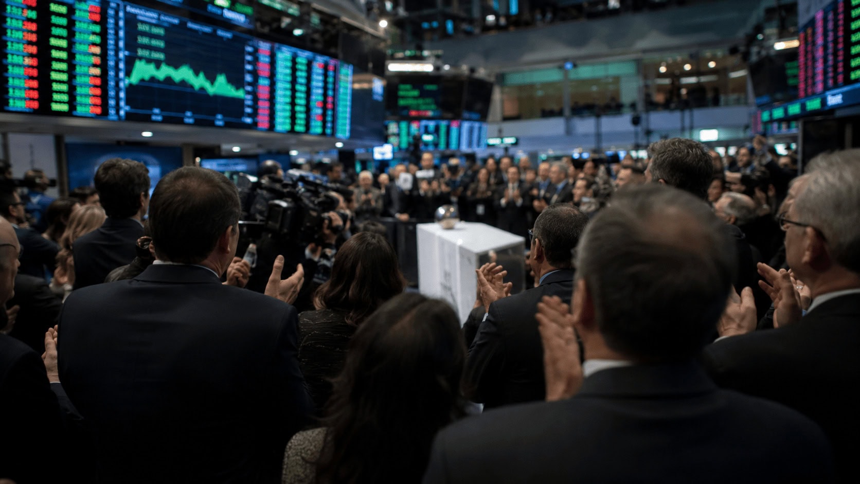 A crowd of people applauding around a podium on a busy stock-exchange trading floor with glowing ticker boards behind them.