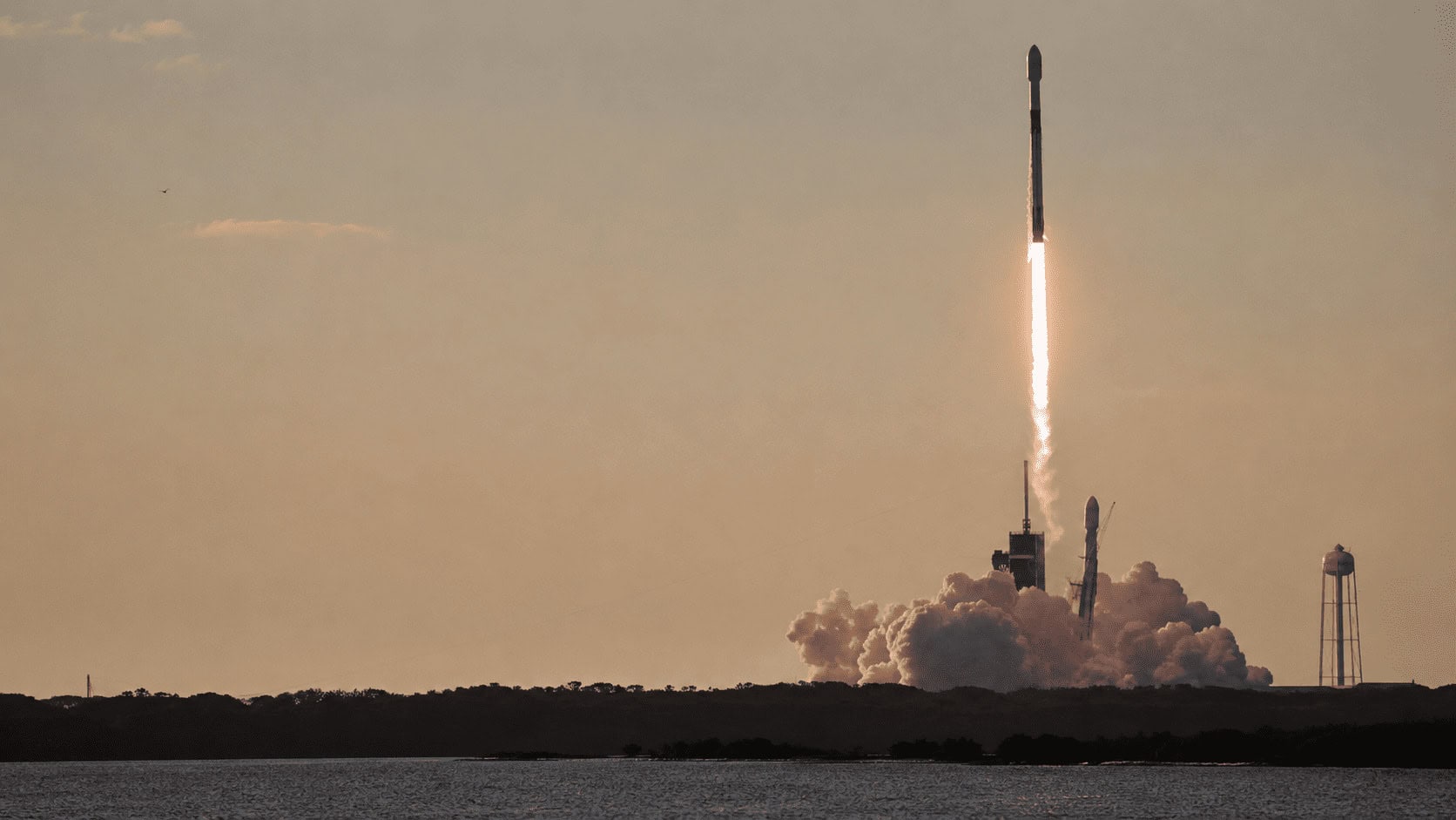 A SpaceX rocket lifting off from a launch pad at dawn, surrounded by exhaust plume and morning haze.