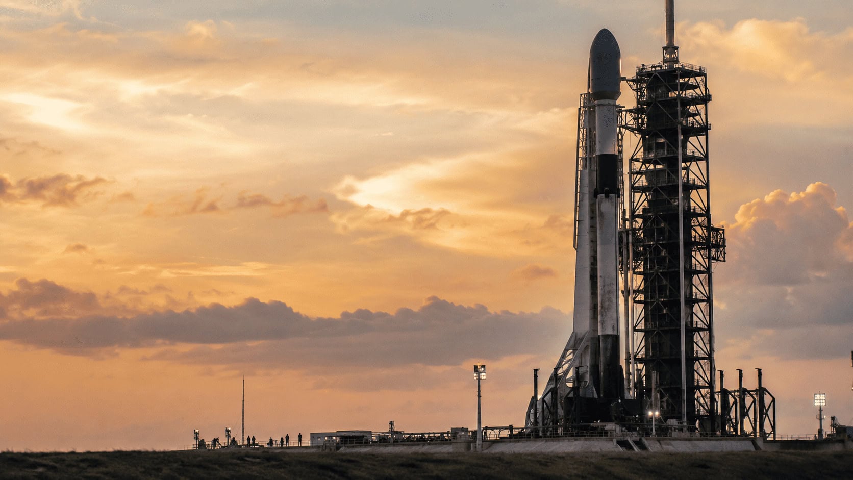 A space rocket stands on a launch pad against an open sky at golden hour, with small ground crew silhouettes in the distance.