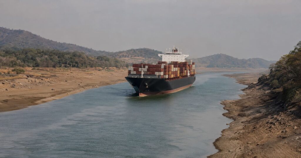 Container ship transiting a drought-lowered shipping canal under hazy heat, illustrating El Niño supply chain disruption.