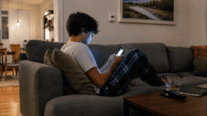 A teenager sits alone on a sofa at night, face lit by the glow of a smartphone held close.
