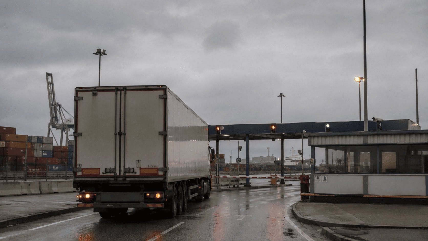 A refrigerated freight truck waiting at a UK port customs checkpoint at dawn with container stacks behind it.