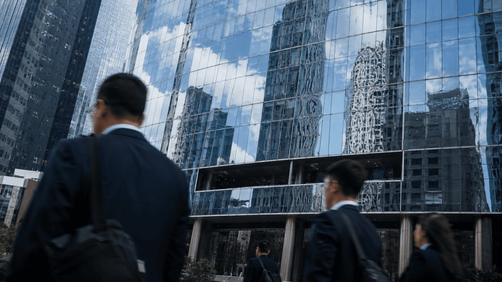 Glass facade of an investment bank office tower reflecting nearby skyscrapers, with blurred pedestrians walking past at street level.