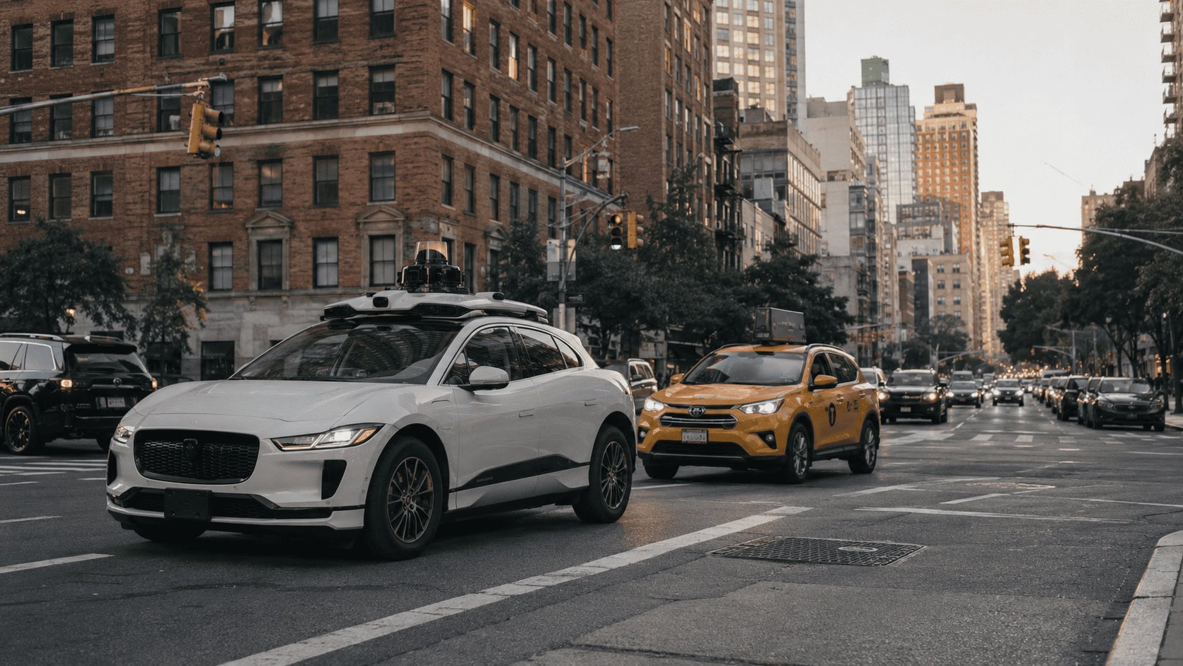 A white self-driving robotaxi waits at a city intersection next to a yellow taxi in evening traffic.