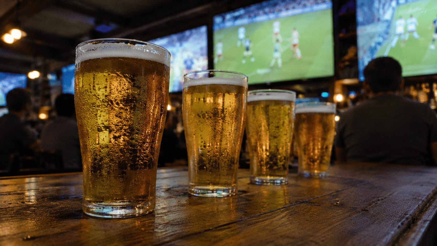 Pint glasses of light lager on a wooden pub bar with televisions showing a soccer match in the warmly lit background.