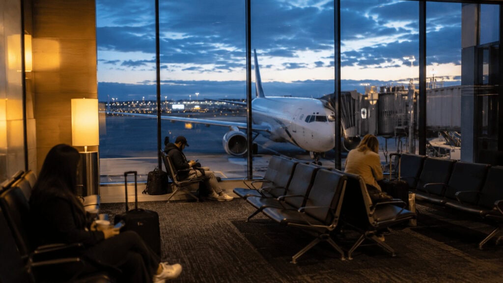 A sparsely occupied airport departure gate at dusk with a parked airplane visible through the windows and a few travelers seated with luggage.